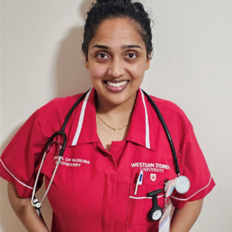 Shiralee Chand standing against a white wall smiling at the camera wearing a red and white nursing uniform with a stethoscope hanging around her neck